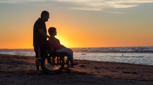 A man behind a woman on a wheelchair looking into the horizon as the sun sets
