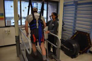 Man supported with harness walks on a treadmill with a female clinician standing beside him