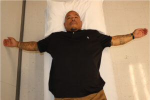 Man supine in bilateral stretch of anterior shoulder structures with “open book” stretch.