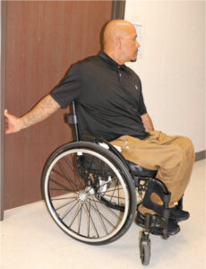 Man seated in wheelchair stretching front of one shoulder using a doorway.