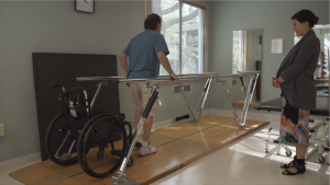 A person walking between parallel bars with his wheelchair behind him. A heathcare provider on the side watches him.