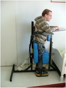 Photograph of a person using a standing frame with cushions behind and in front of the legs to help align them. The person is resting their arms on a small desk that is attached to the frame.