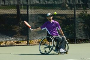 A man in a wheelchair swinging his racquet to hit a tennis ball.